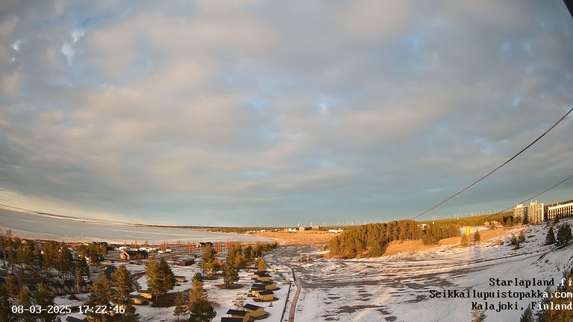 A partly cloudy sunset over a snow-covered landscape with small cabins, a frozen lake, and wind turbines in the distance.