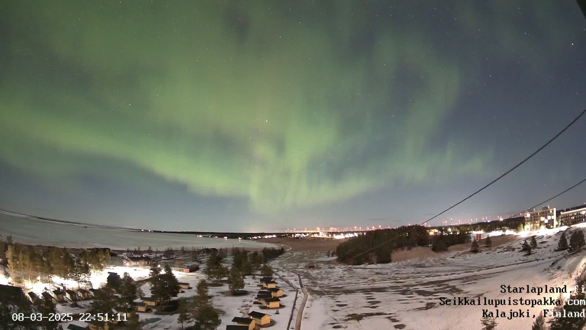 A vibrant green aurora borealis illuminates a snowy landscape at night, with a body of water and small buildings visible in the foreground.