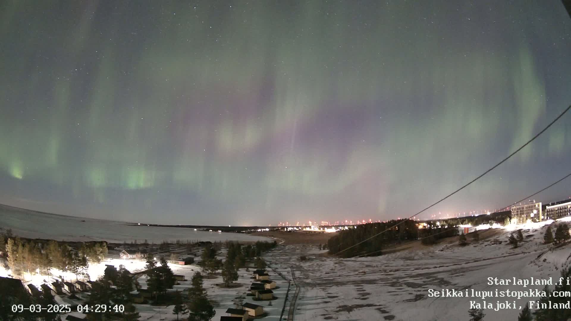 A vibrant aurora borealis illuminates a snowy landscape at night, with a distant town faintly visible in the background.