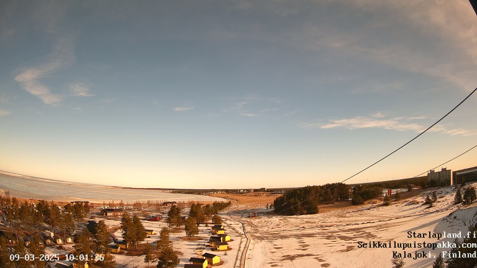 A mostly clear, twilight sky overlooks a snow-dusted landscape with a frozen lake and small, yellow buildings near a sparsely wooded area.