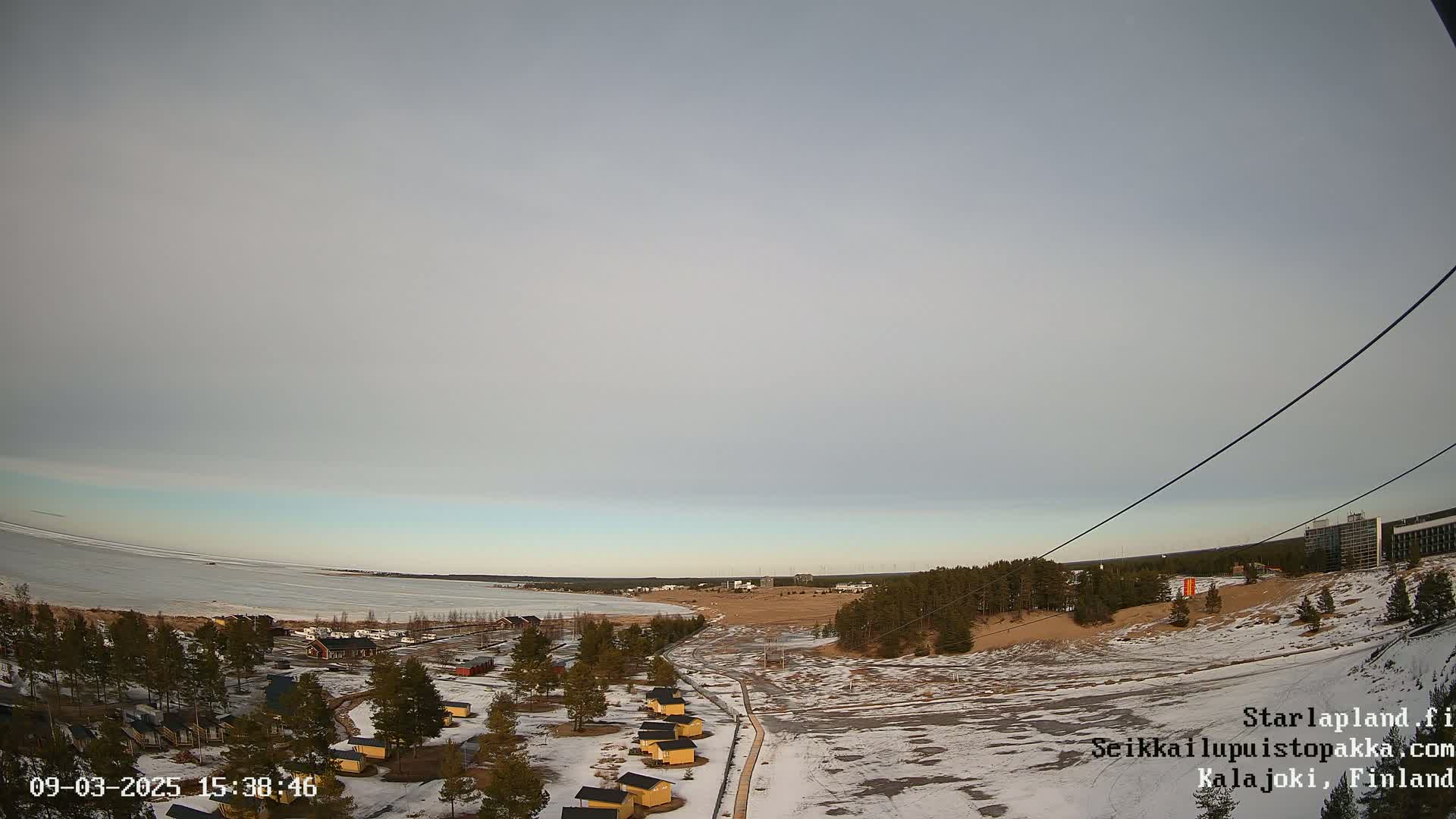 A partly cloudy, winter day overlooks a snow-covered landscape with a frozen lake, small cabins, and a larger building in the distance.