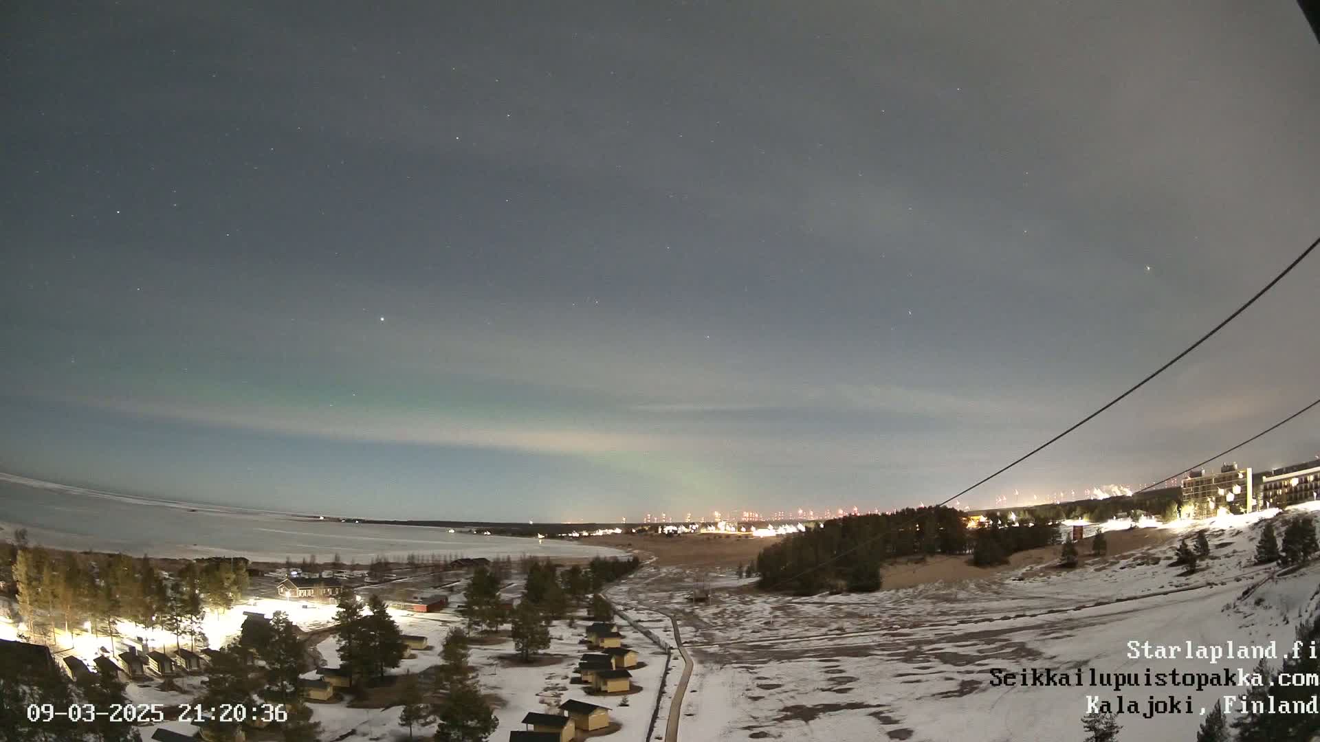 A nighttime view of a snow-covered landscape near a frozen lake, under a mostly clear sky displaying a faint aurora borealis.