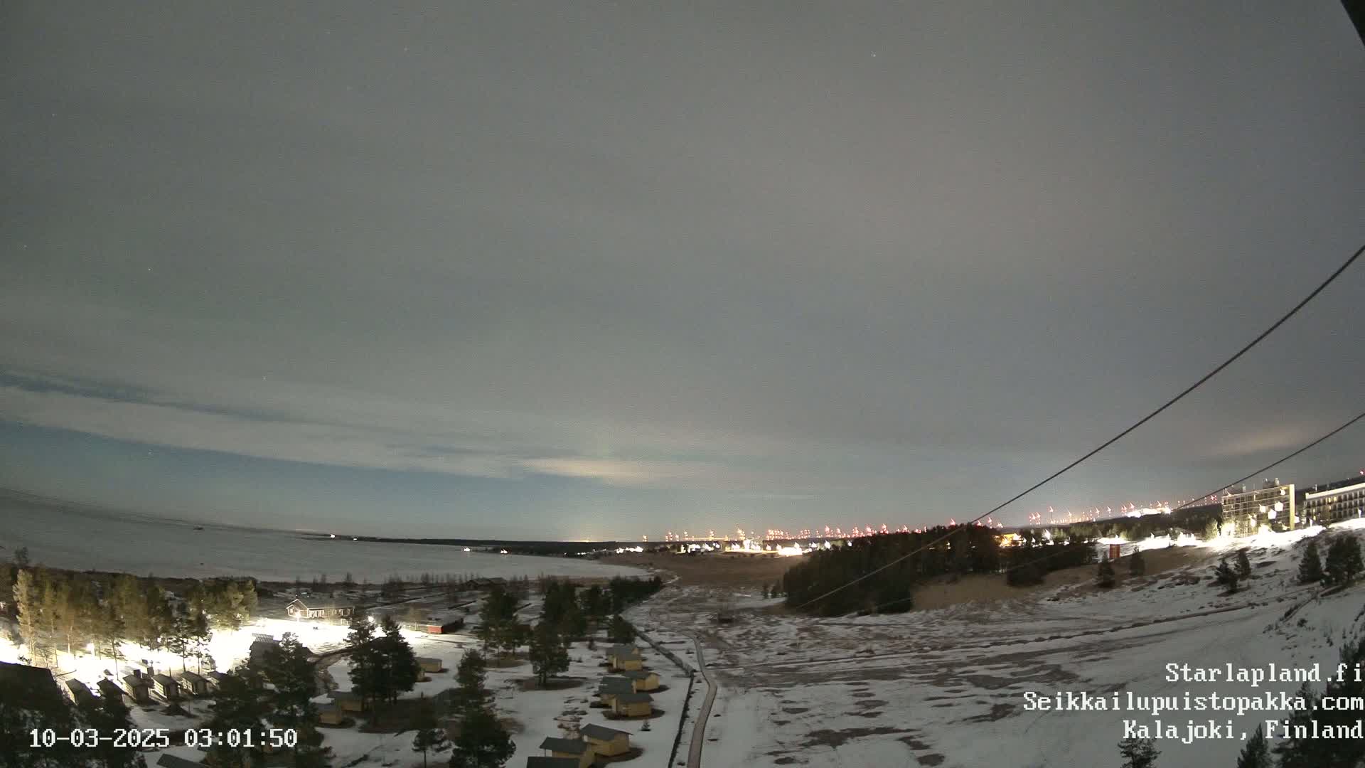 A nighttime, snow-covered landscape overlooks a frozen body of water and a distant town illuminated under a cloudy sky.