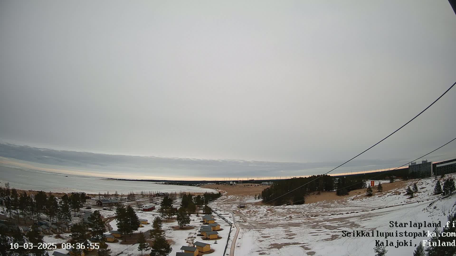 A mostly cloudy sky overlooks a snow-covered landscape with small cabins, a frozen body of water, and a distant building.