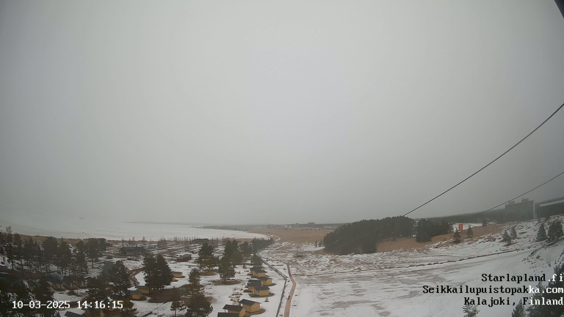A snow-covered landscape with small cabins and a partially frozen body of water under an overcast sky.