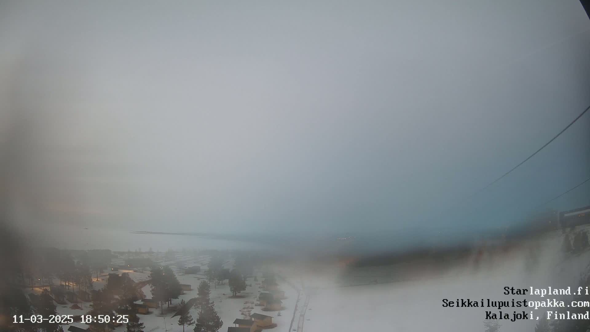 A hazy, overcast view shows a snow-covered landscape with small buildings and a body of water in the distance.