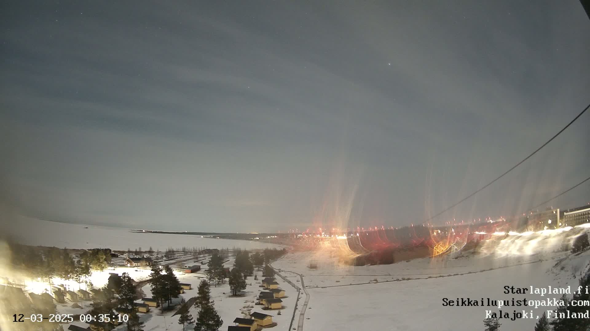 A snow-covered landscape at night, illuminated by lights from buildings and what appear to be light trails from vehicles, under a mostly clear sky.