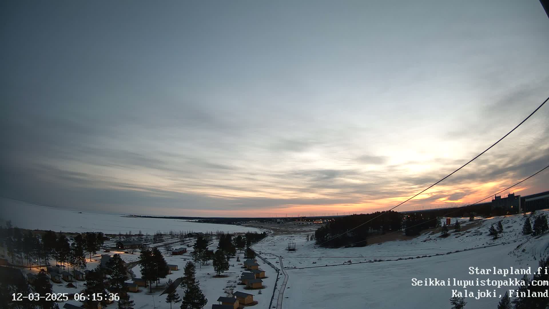 A snow-covered landscape at sunset, with a cluster of small cabins visible in the foreground and a distant horizon featuring a line of trees and wind turbines under a mostly clear sky.
