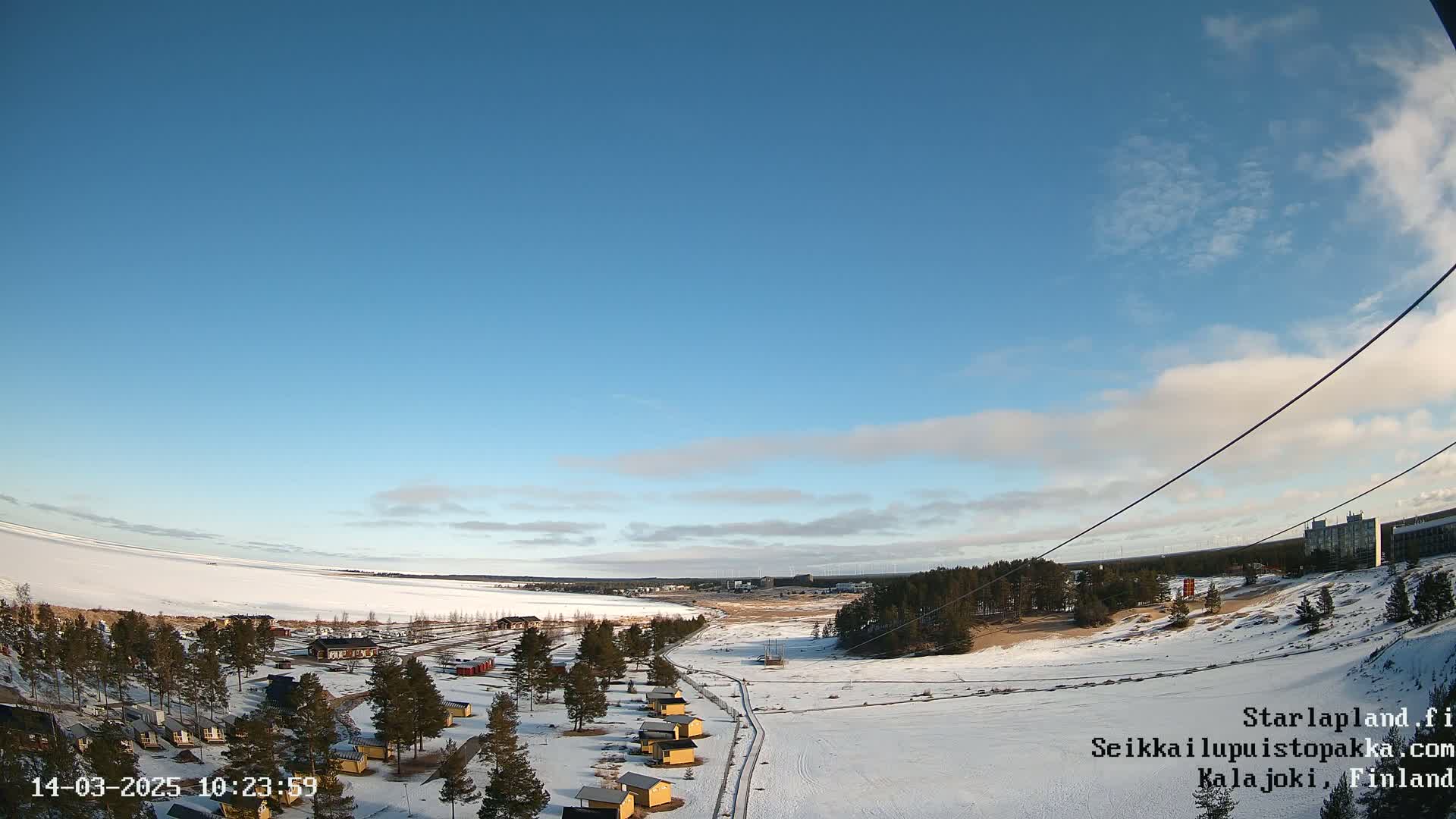 A snow-covered landscape with small buildings and a frozen lake under a mostly clear blue sky.