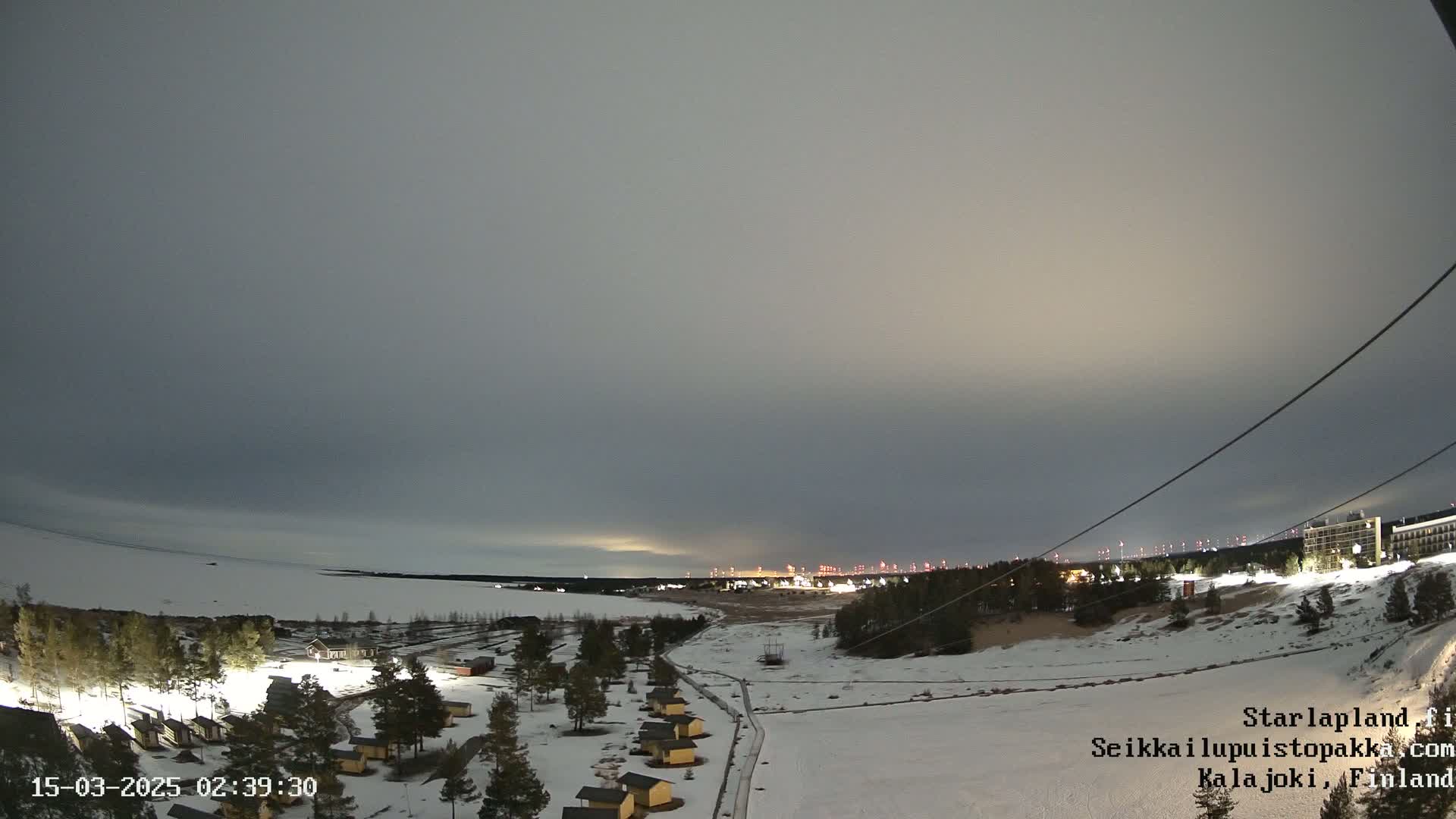 A snow-covered landscape at twilight, with a scattering of small buildings and a distant town faintly illuminated under a mostly cloudy sky.