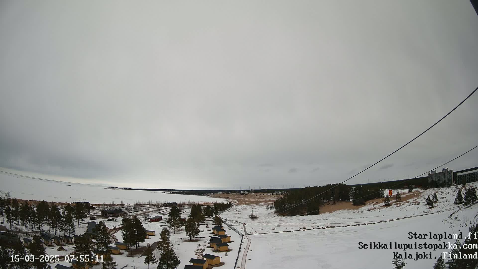 A snow-covered landscape with small cabins, a frozen lake, and a mostly cloudy sky is viewed from above.