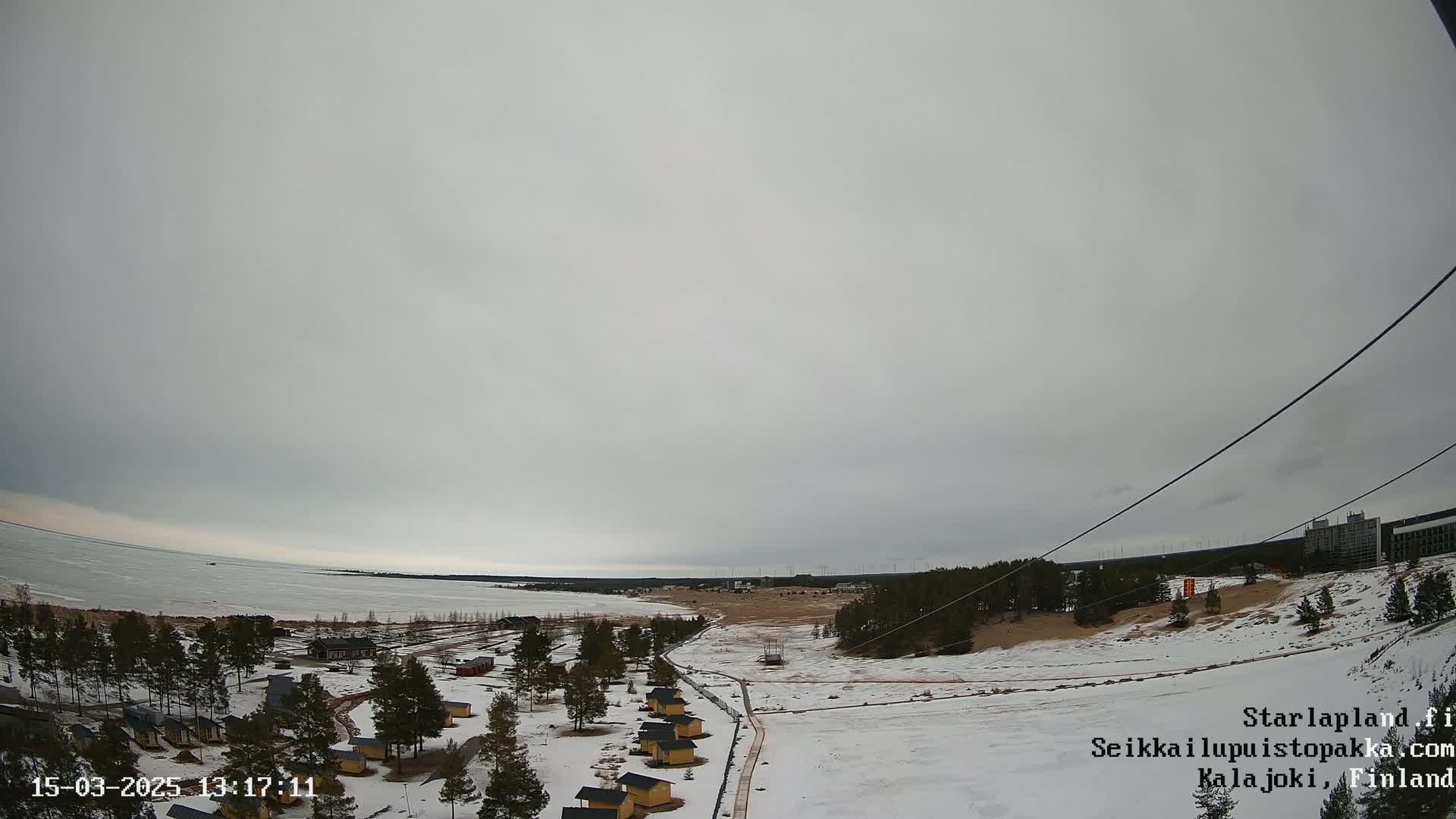 A snow-covered landscape with small buildings and a partially frozen body of water under an overcast sky is visible.