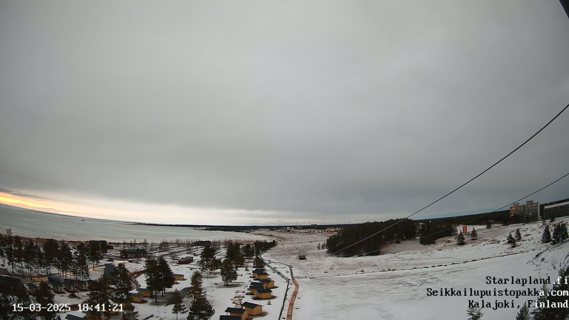 A snow-covered landscape with small cabins near a frozen lake under an overcast sky.