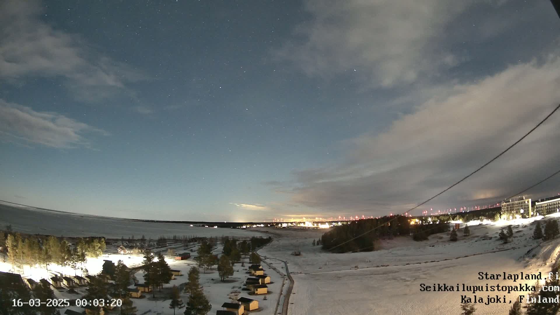 A nighttime, snow-covered landscape with a few small buildings lit up under a partly cloudy sky dotted with stars.