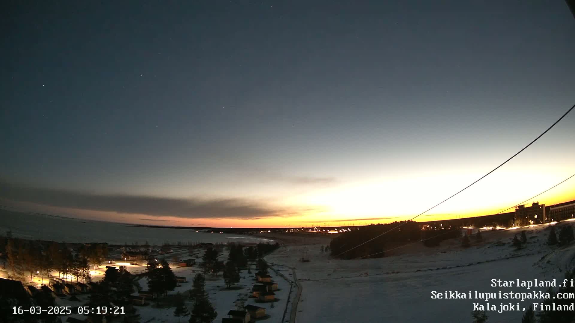 A twilight sunset over a snow-covered landscape with a body of water and small buildings illuminated by lights.