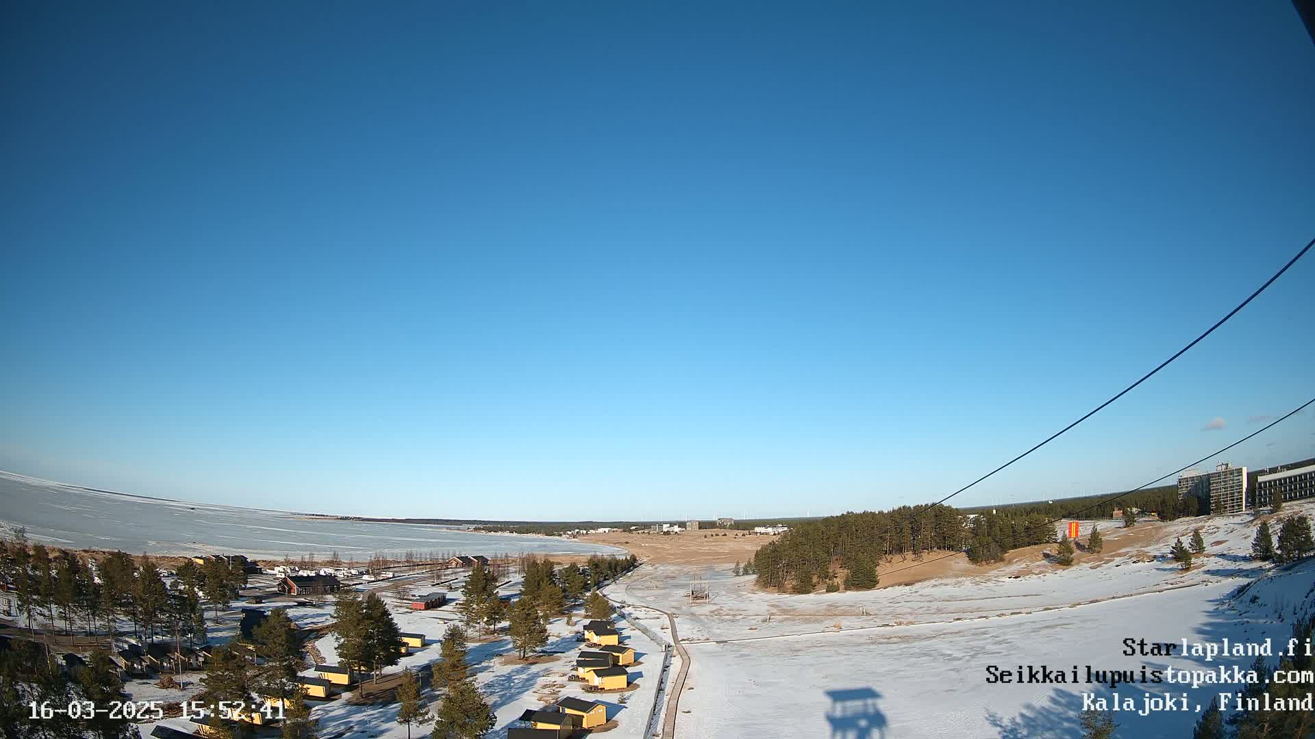 A snow-covered landscape with small buildings and a frozen body of water stretches under a clear blue sky.