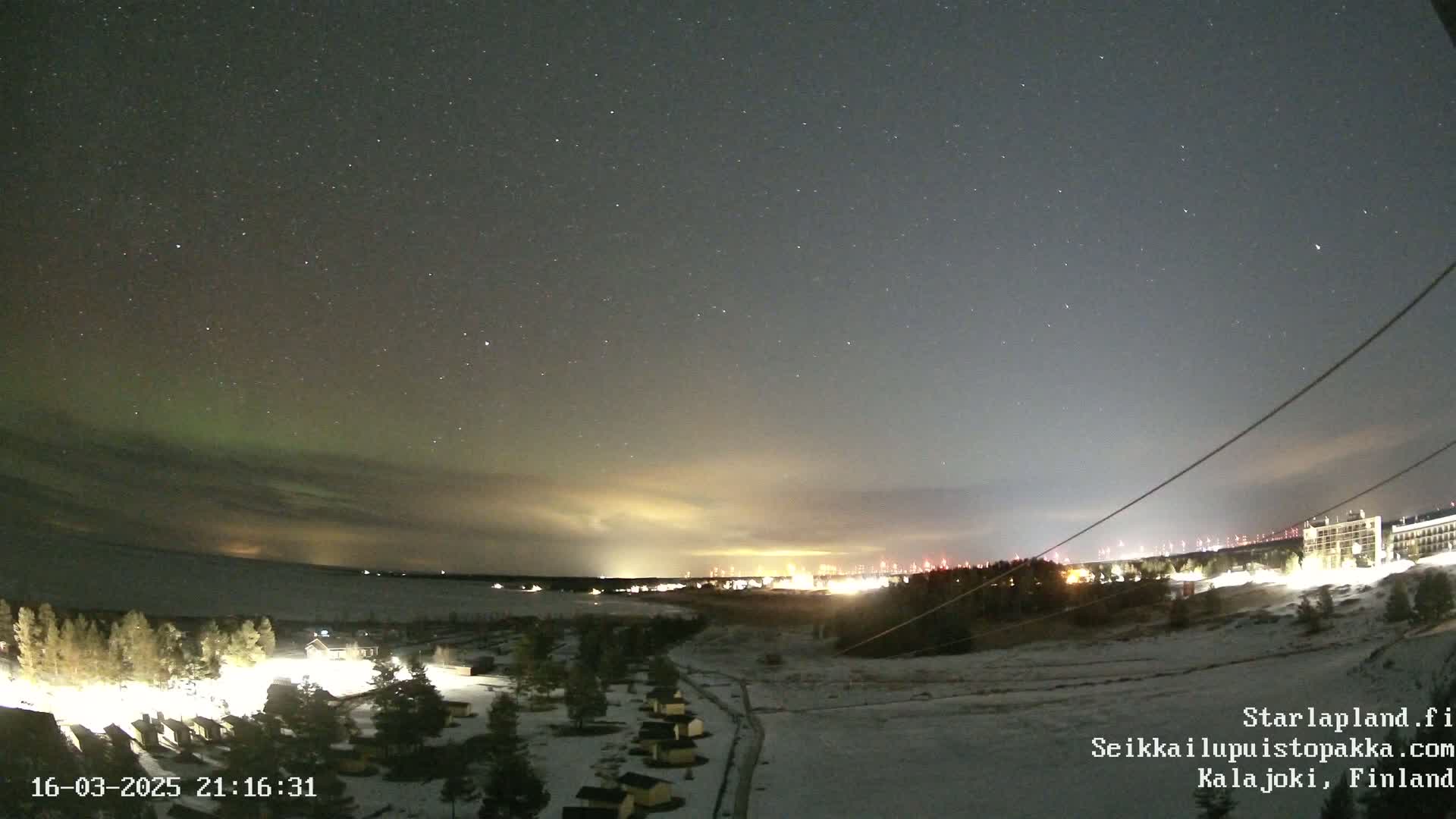 A nighttime landscape shows a faint aurora borealis in the sky above a snow-covered area with small cabins and distant city lights.