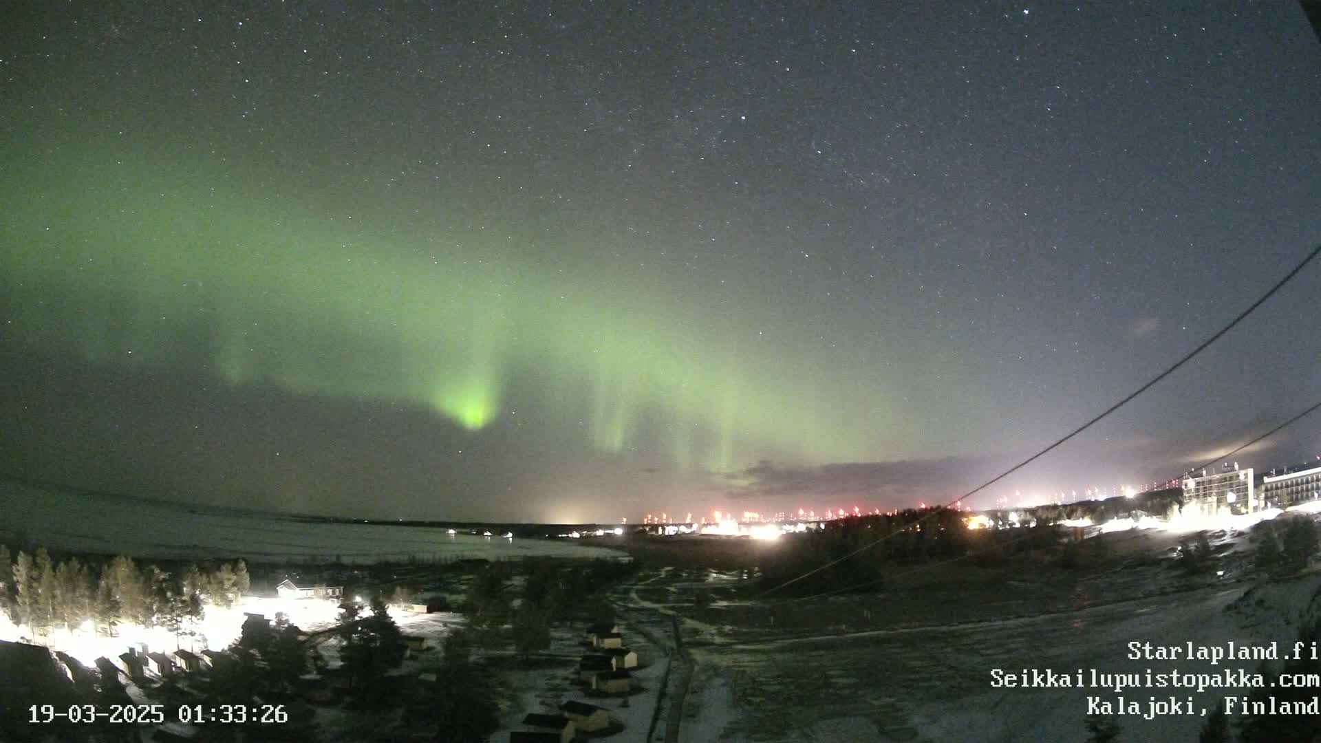 A vibrant green aurora borealis illuminates a clear night sky over a snow-covered landscape with a distant town and scattered buildings.
