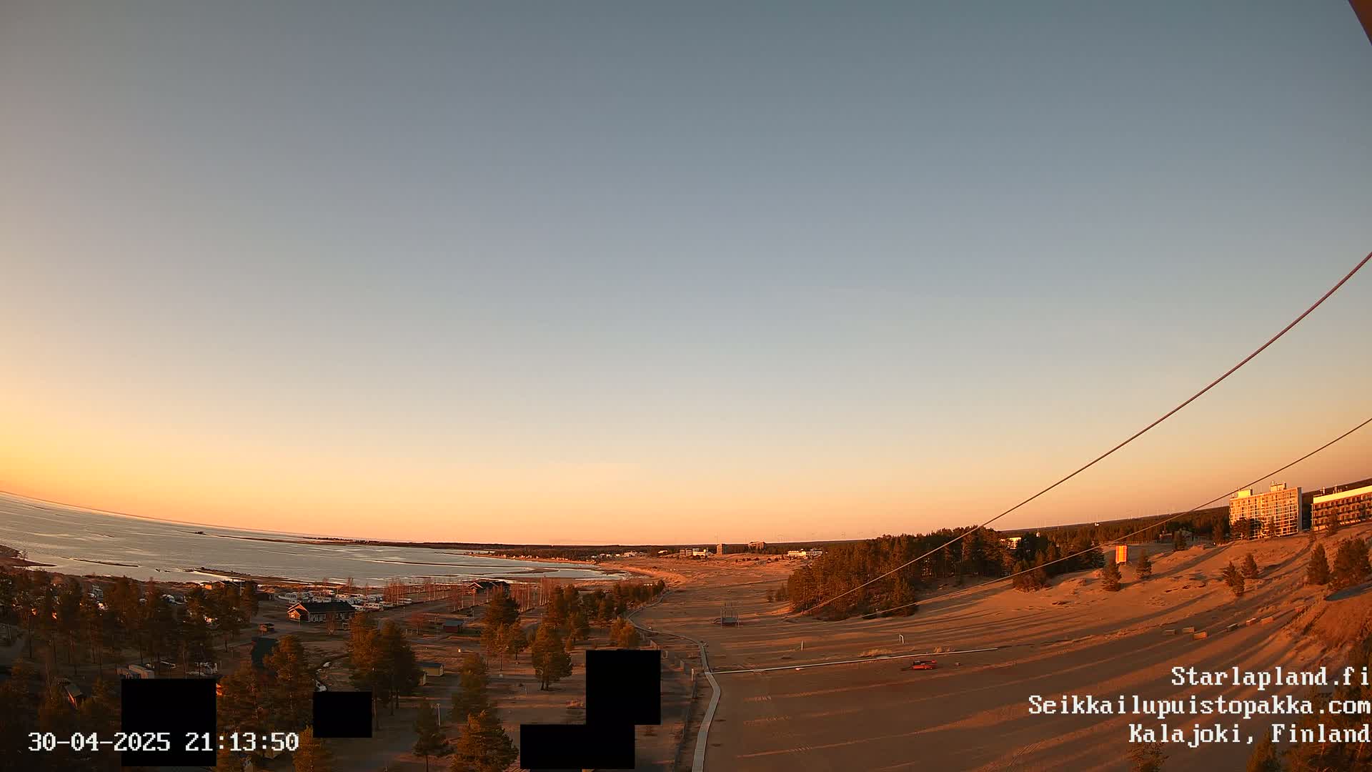 A sunset over a sandy beach and calm body of water, with sparse trees and buildings visible in the distance.