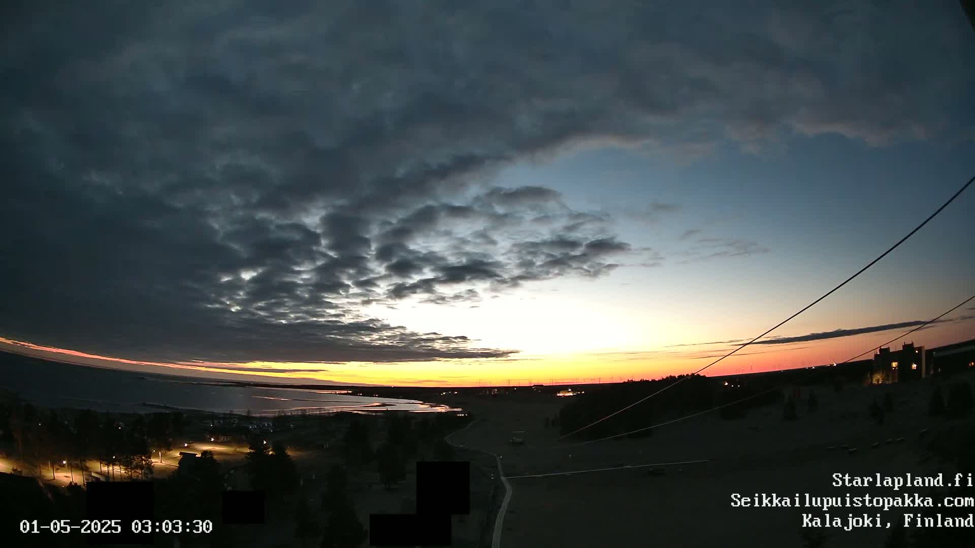 A dark, cloudy sunset over a calm body of water, with a few lights visible on the shore and in the distance.
