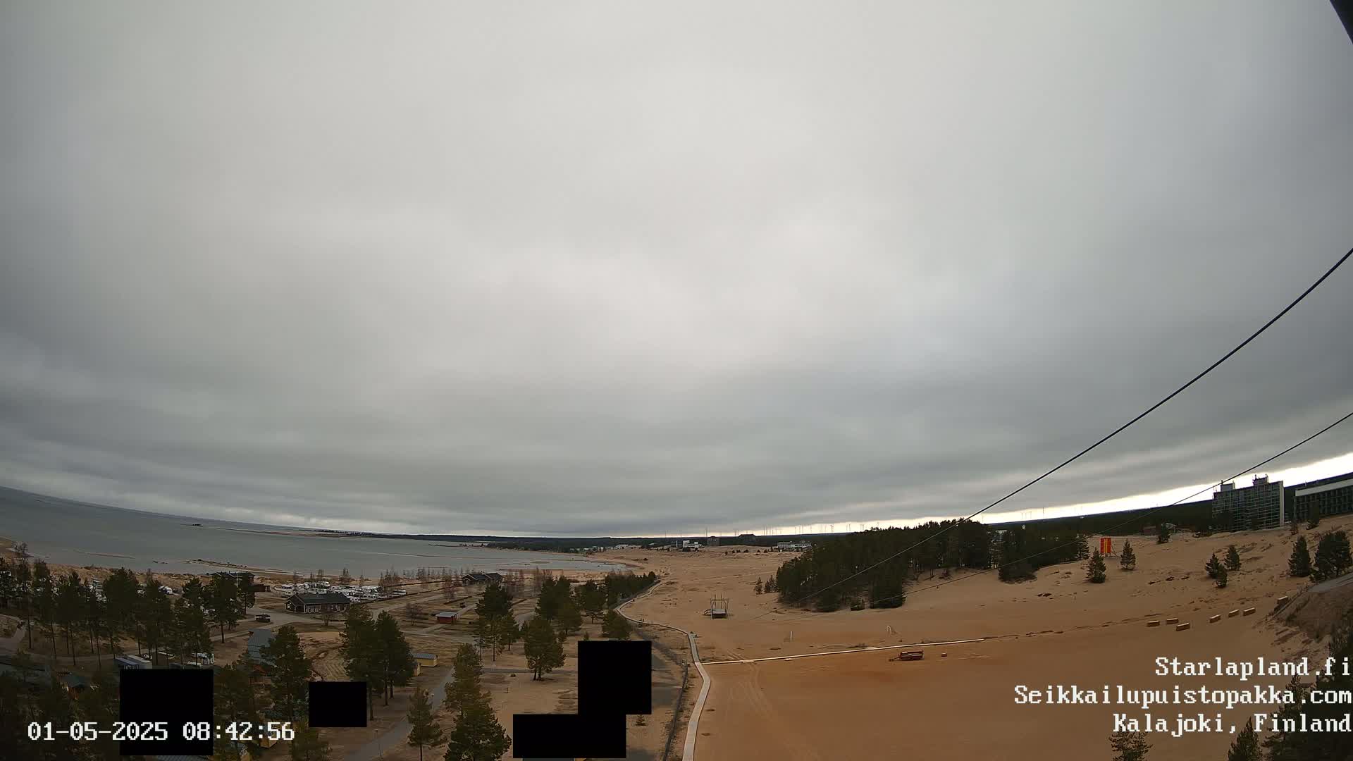A wide, overcast shot shows a sandy area near a body of water, with trees, buildings, and a zip line visible in the distance.