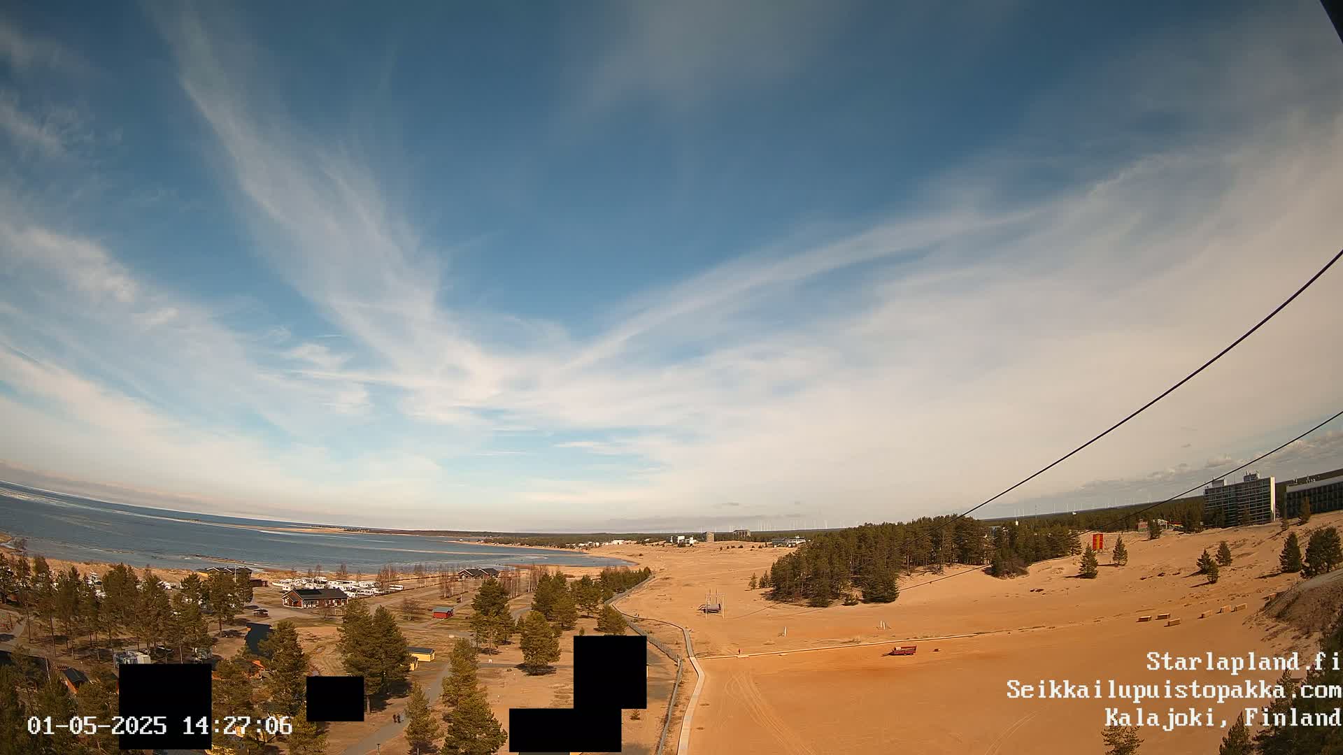 A mostly sunny day with wispy clouds over a sandy landscape near a body of water, featuring a sparse forest and some buildings in the distance.
