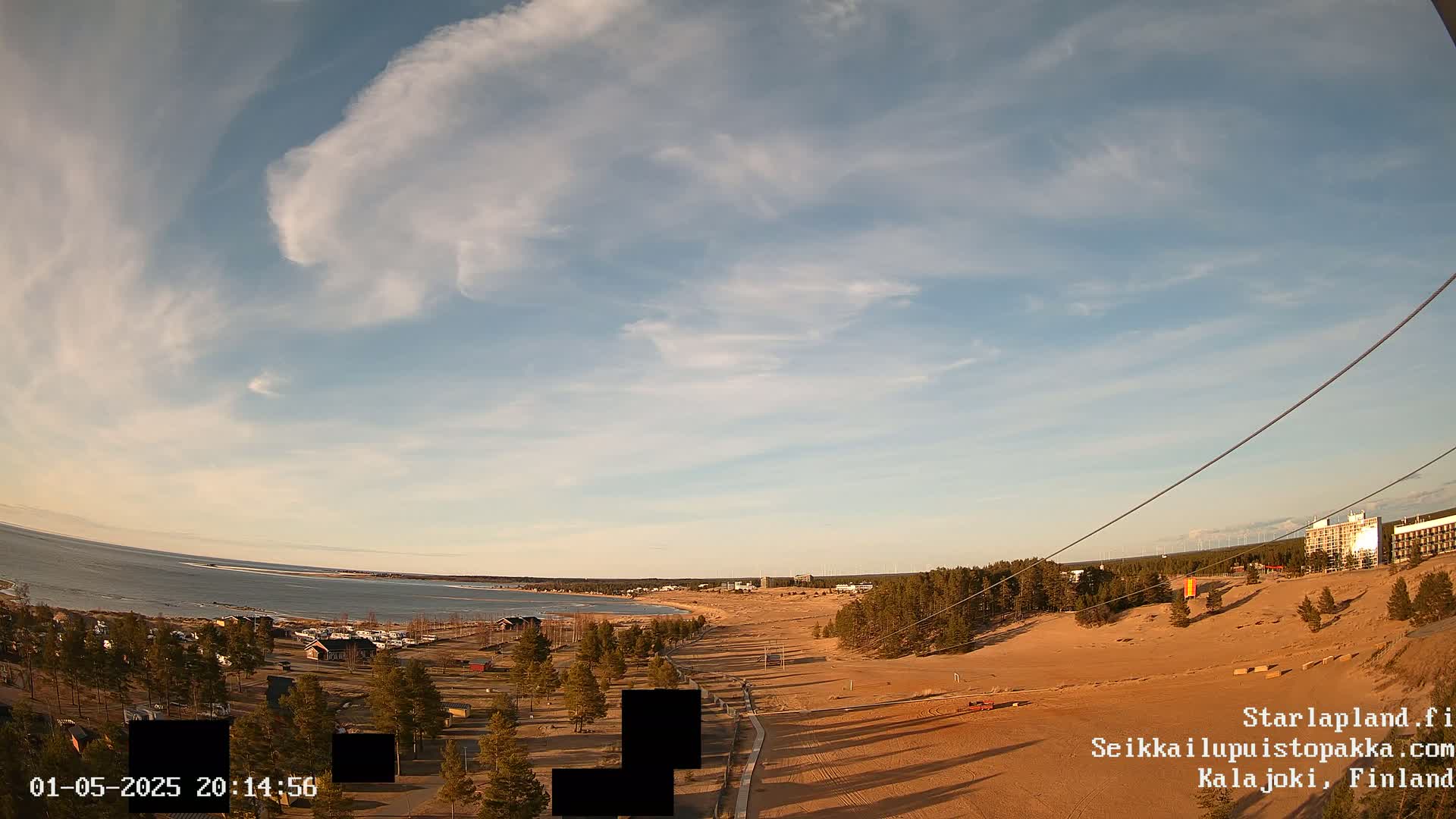 A partly cloudy sky overlooks a sandy beach, a calm body of water, and a sparsely wooded area with buildings in the distance.