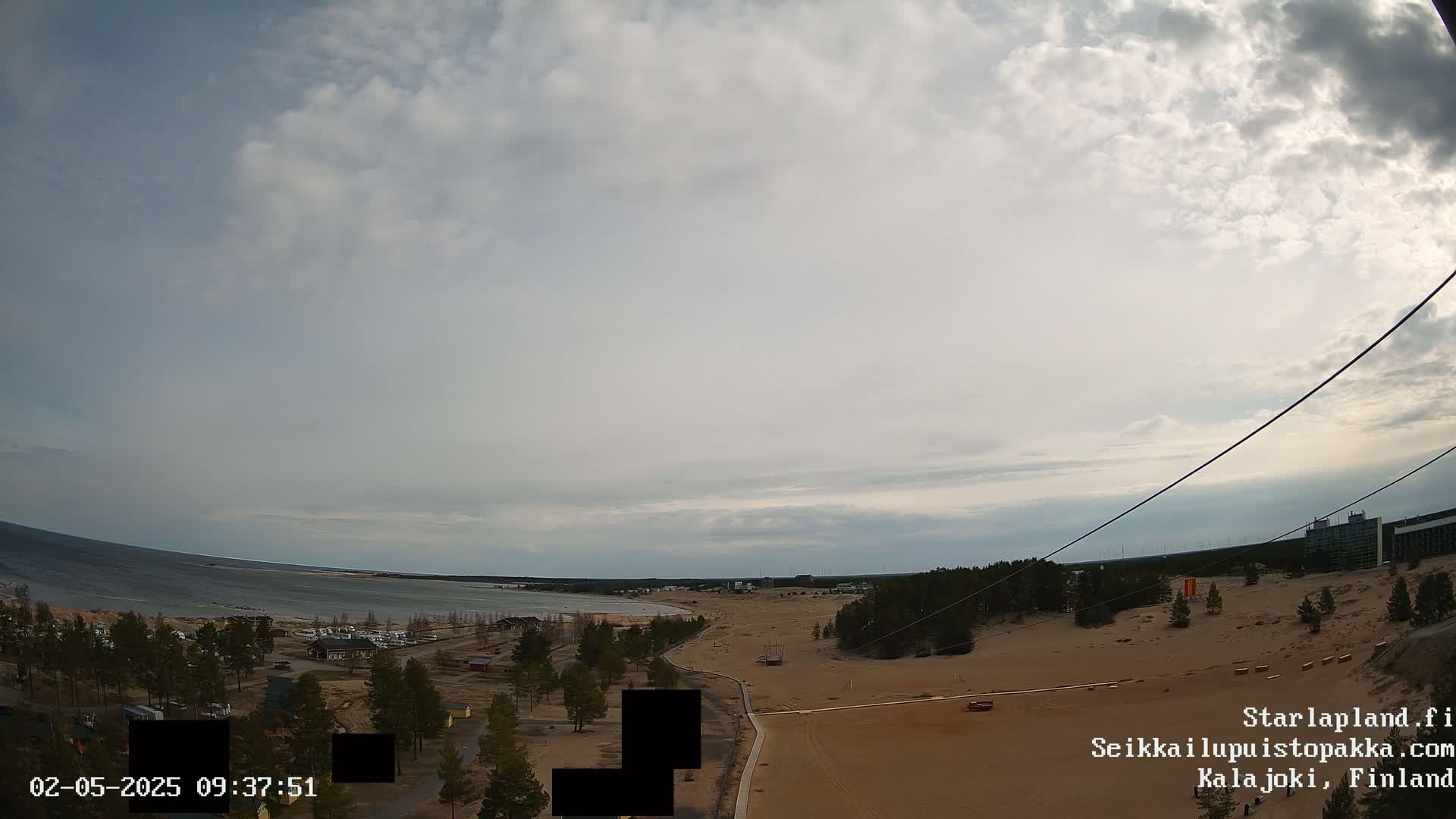 An elevated view shows a sandy landscape with sparse trees, a calm body of water in the distance, and a cloudy sky.