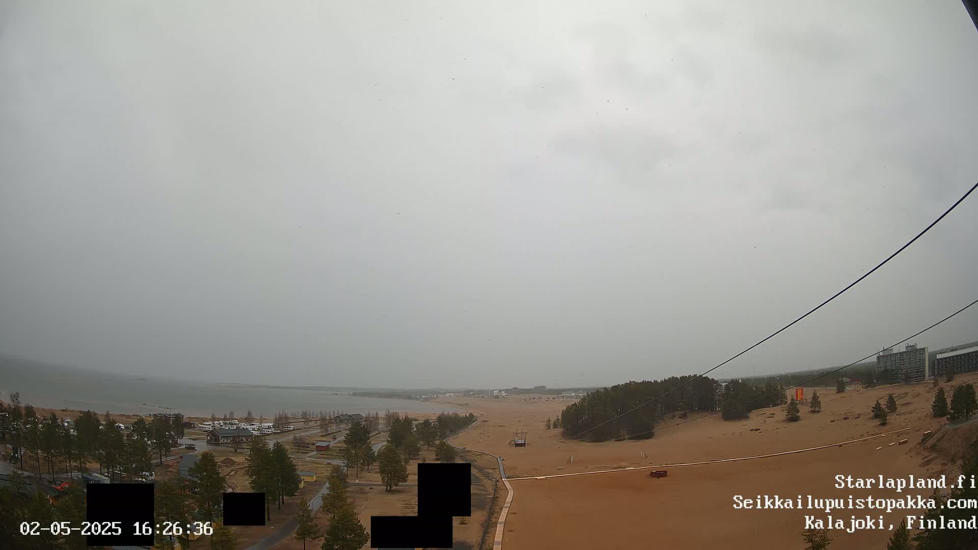 A wide, overcast view shows a sandy beach and calm body of water, with sparse trees and buildings visible in the distance.