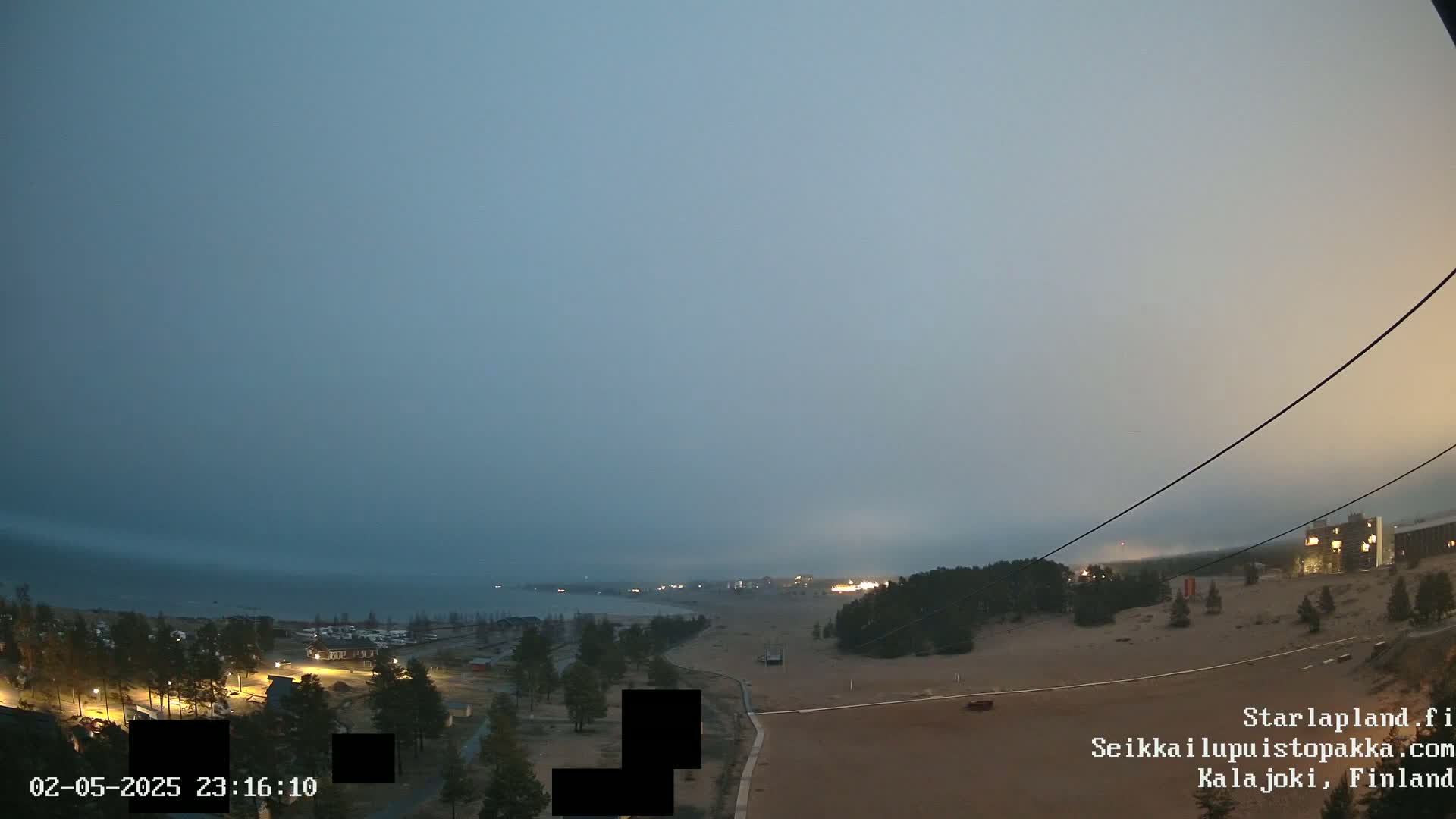An overcast evening scene shows a body of water in the distance, a sandy area in the foreground with sparse trees and buildings, and power lines stretching across the image.