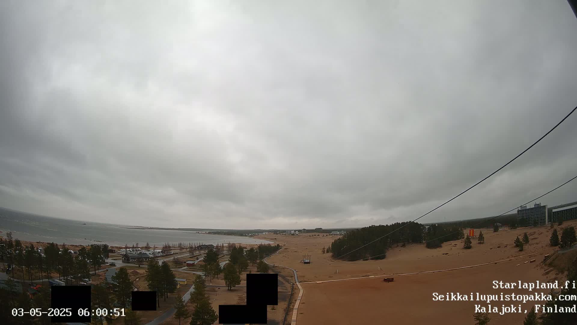 An overcast sky overlooks a sandy landscape with sparse vegetation, a body of water, and buildings in the distance.