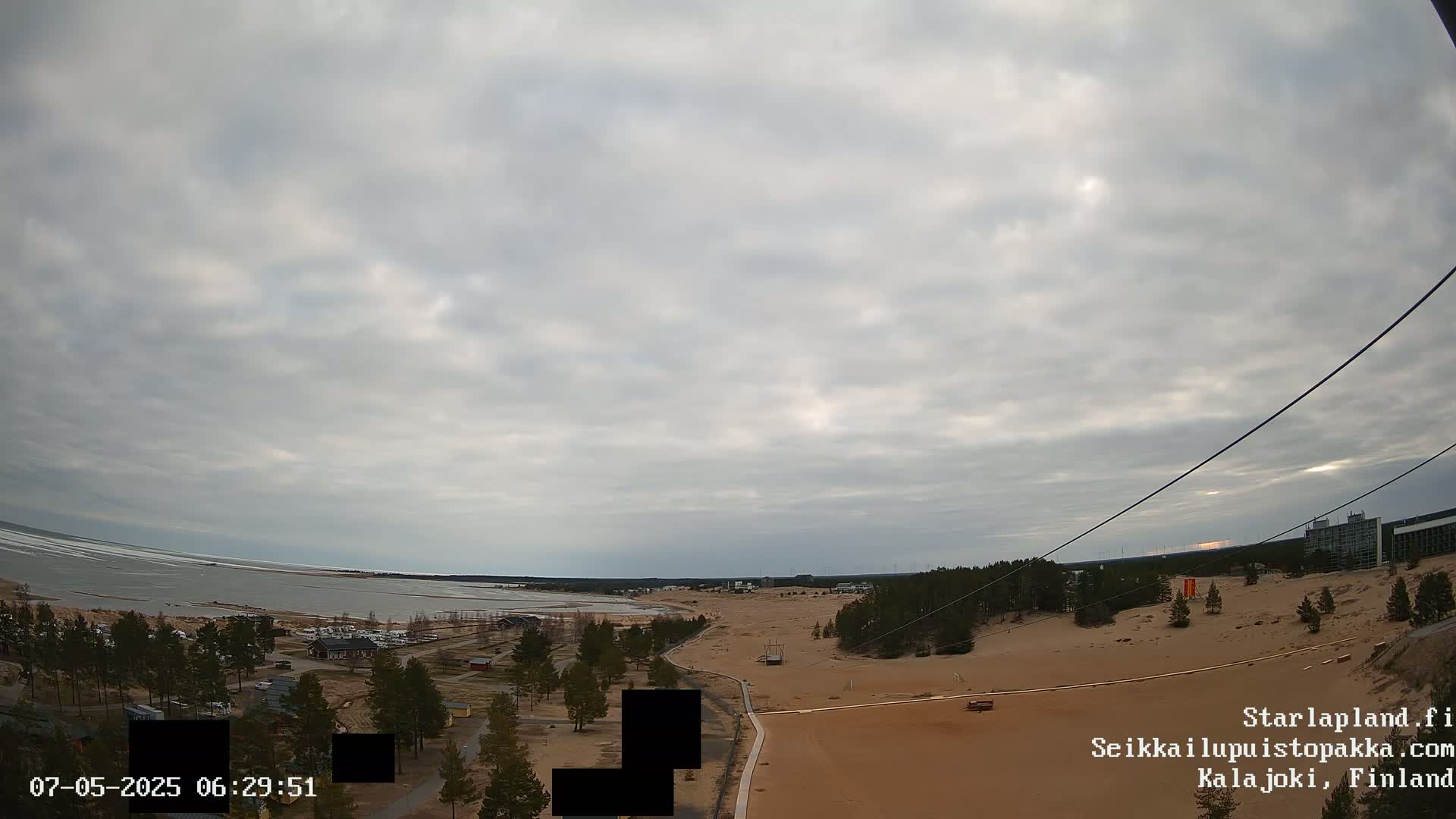 A mostly cloudy sky overlooks a sandy landscape with a body of water and some buildings in the distance.