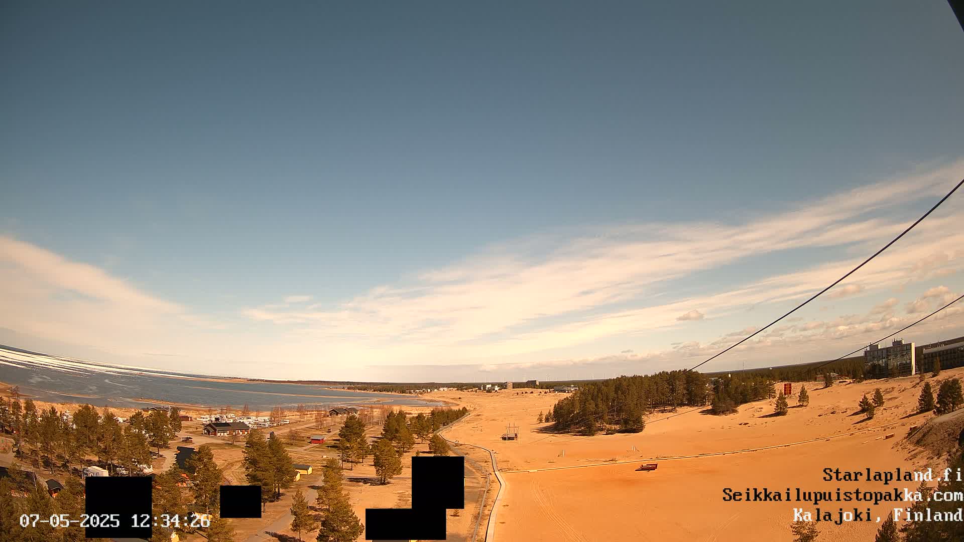 An aerial view shows a sandy landscape next to a body of water under a mostly clear, bright sky.
