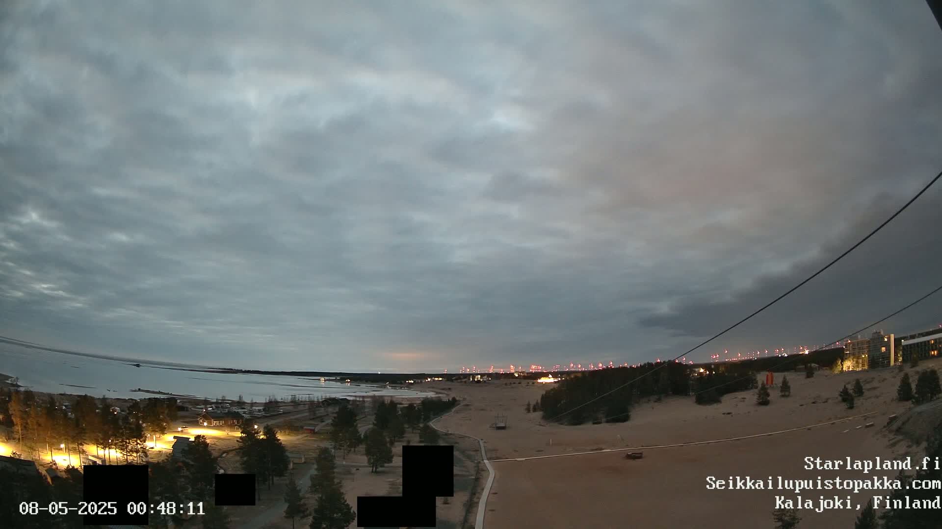 A wide, high-angle view shows a mostly cloudy sky over a sandy landscape with sparse trees, a body of water in the distance, and a few distant lights at dusk.