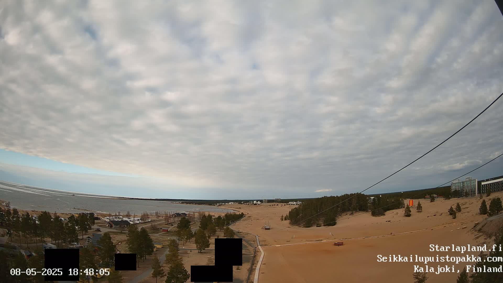 A mostly cloudy sky overlooks a sandy beach and coastline, with a calm body of water visible in the distance.