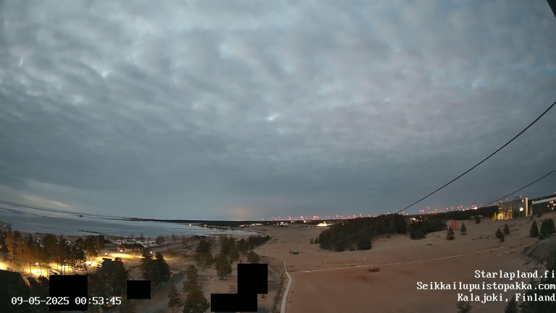 A mostly cloudy twilight sky overlooks a coastal landscape with sparsely vegetated dunes, a body of water, and distant lights.