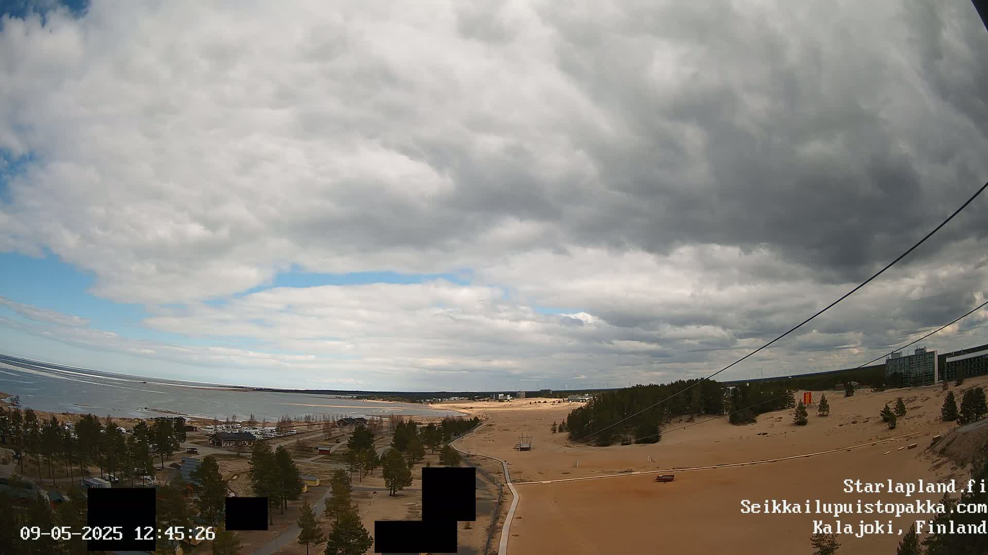 A mostly cloudy sky overlooks a sandy beach and coastline with sparse trees and buildings, a calm body of water visible in the distance.