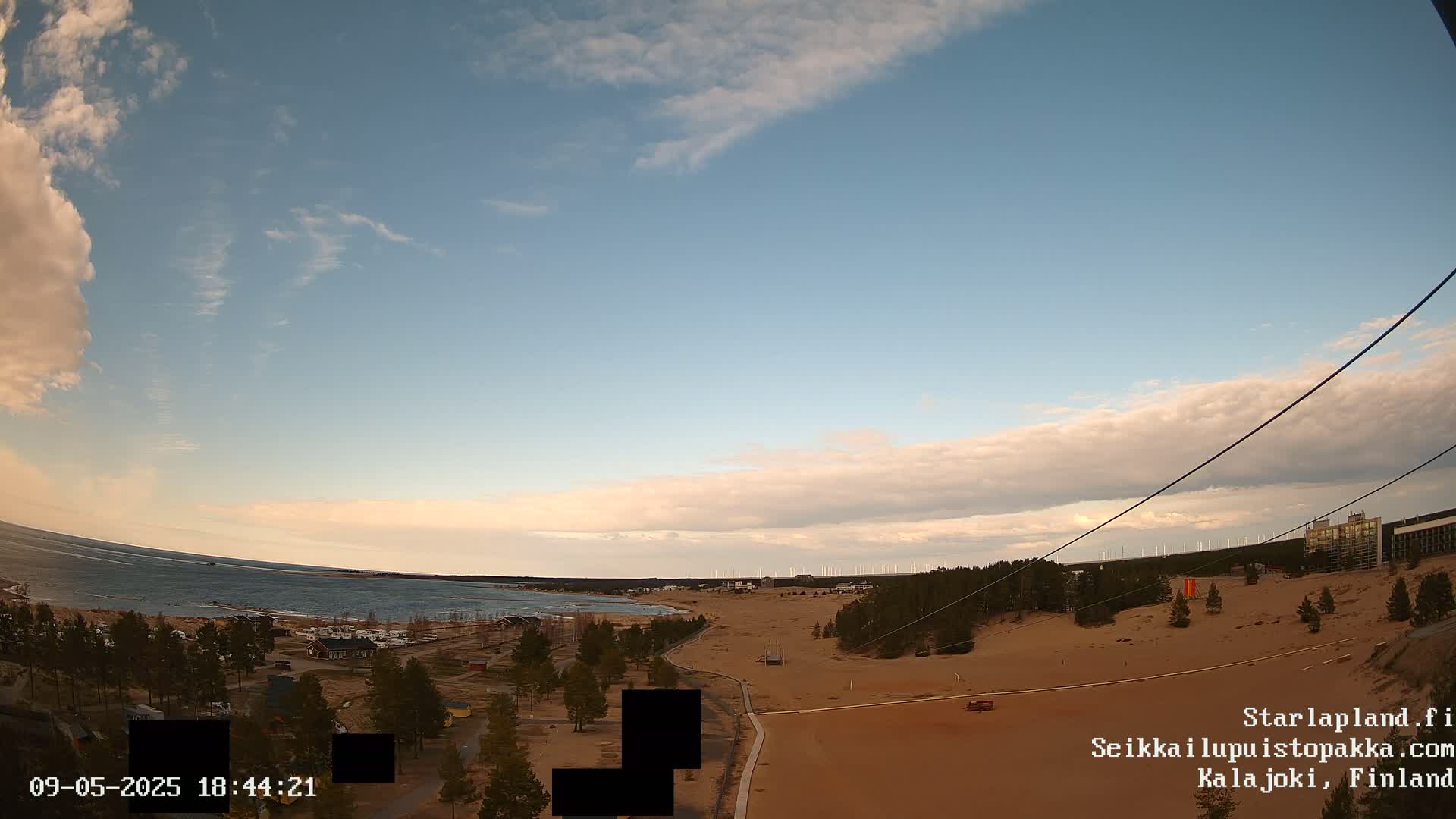 A mostly clear sky with some thin clouds hangs over a sandy landscape near a calm body of water, with distant wind turbines visible on the horizon.