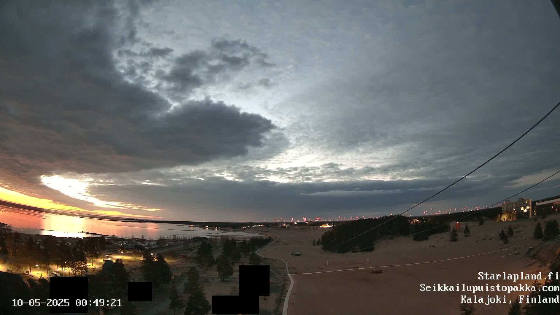 A twilight view over a calm body of water, showing a mostly cloudy sky with hints of sunset colors, and a sandy landscape with sparse vegetation and distant buildings.