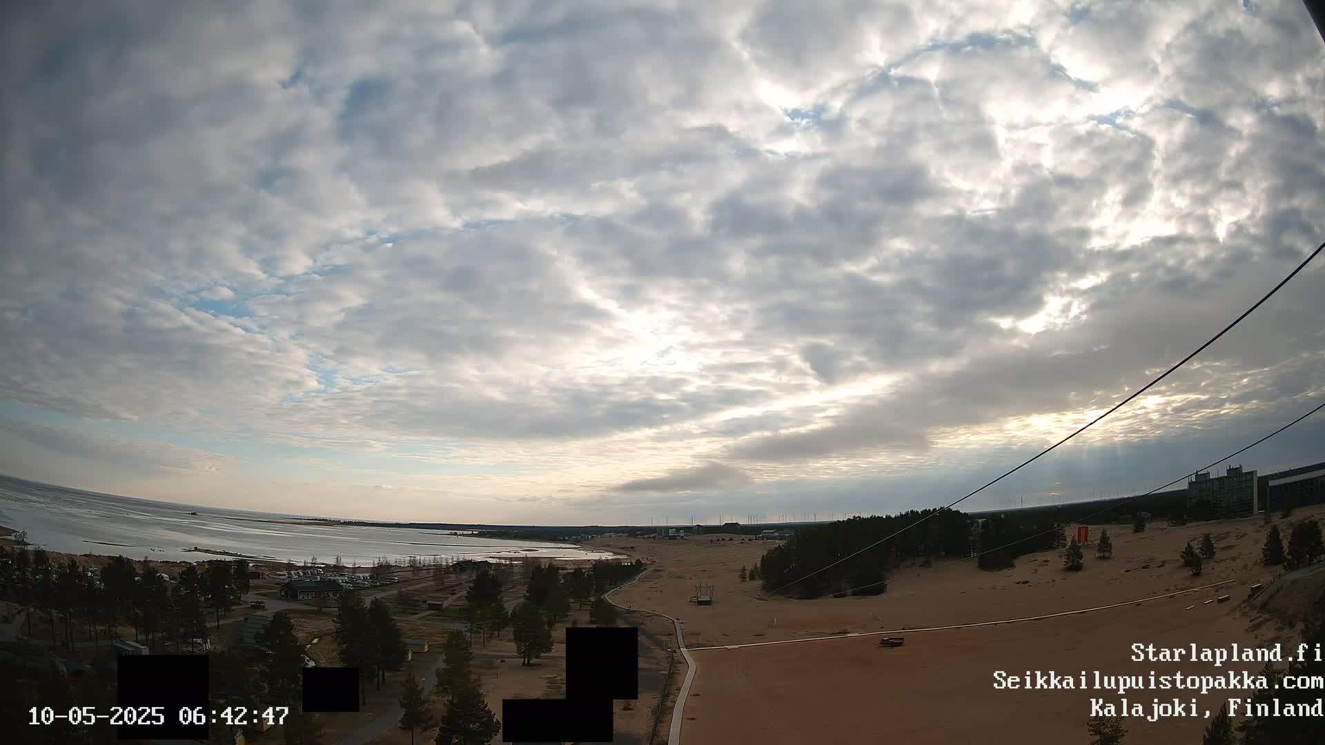 A mostly cloudy sky over a sandy landscape with a body of water in the distance, featuring some trees and buildings.