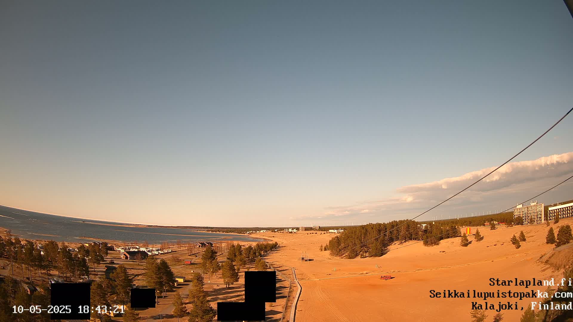 A mostly clear sky overlooks a sandy beach and calm body of water, with sparse vegetation and a few buildings visible in the distance.