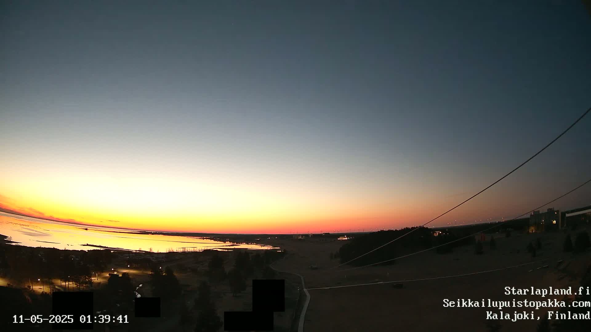 A calm body of water reflects the warm colors of a setting sun under a clear, twilight sky over a flat landscape with sparse vegetation and some buildings.