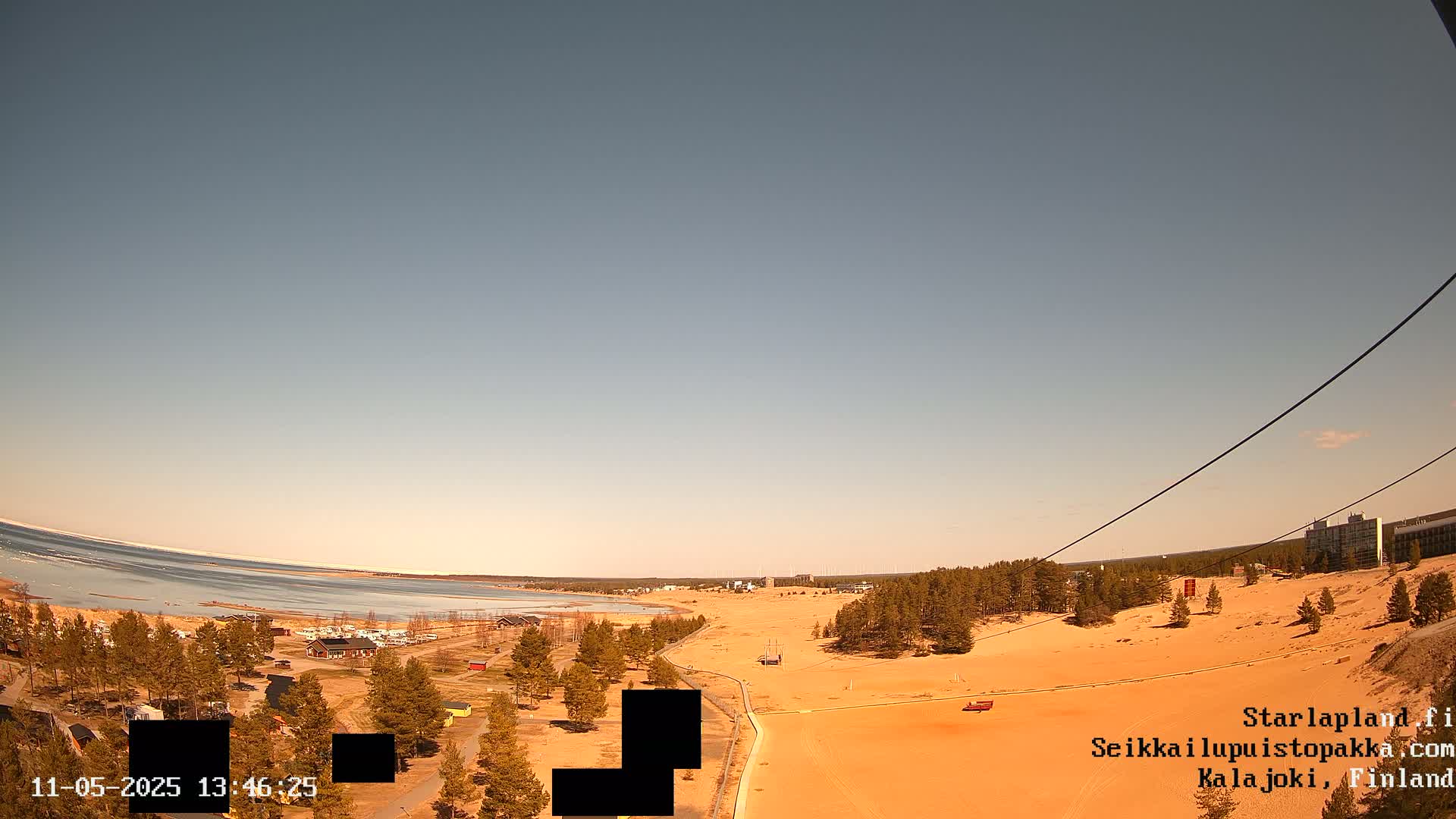 A mostly clear, bright sky overlooks a sandy landscape with sparse vegetation, a body of water with some ice visible in the distance, and a few buildings.