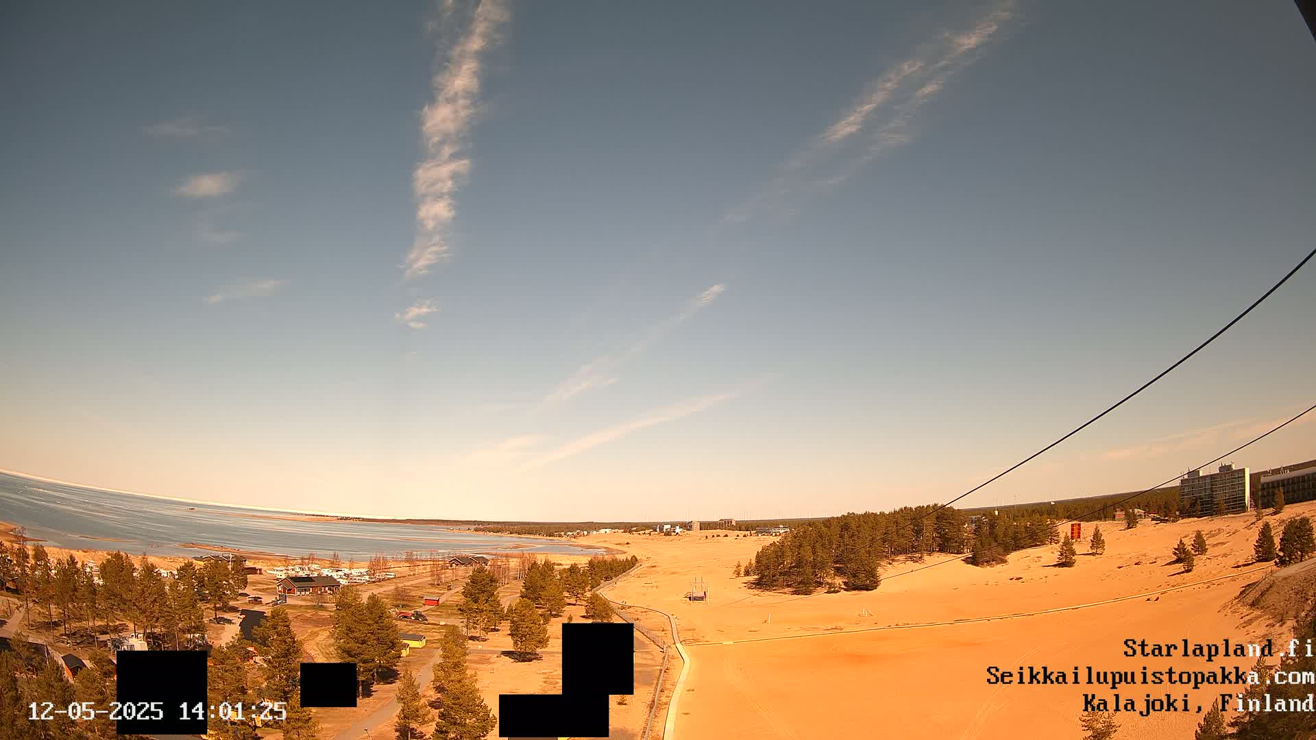 A mostly sunny day reveals a sandy landscape overlooking a partially frozen lake, with a small town and distant buildings visible.