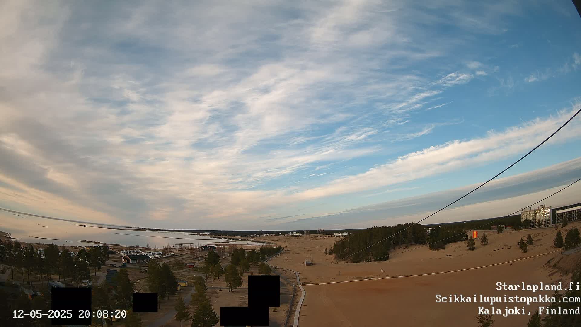 A high-angle view shows a sandy landscape near a body of water under a mostly clear, light blue sky with wispy clouds.