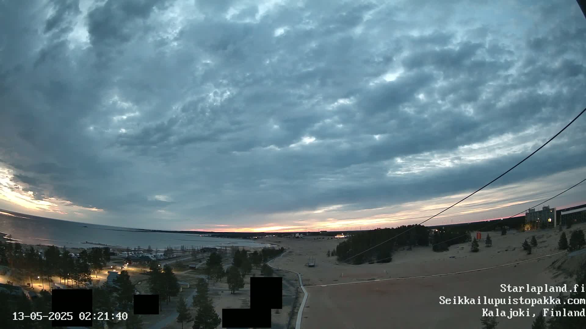 A twilight sunset over a calm body of water, viewed from a high vantage point overlooking a sandy area with sparse vegetation and buildings in the distance, under a mostly cloudy sky.