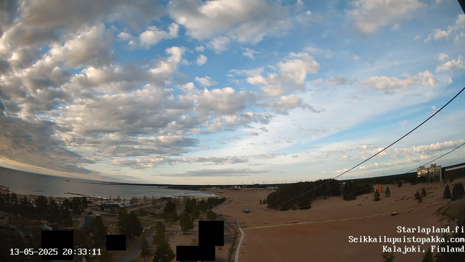 A partly cloudy sky overlooks a sandy landscape with a body of water, trees, and buildings in the distance.