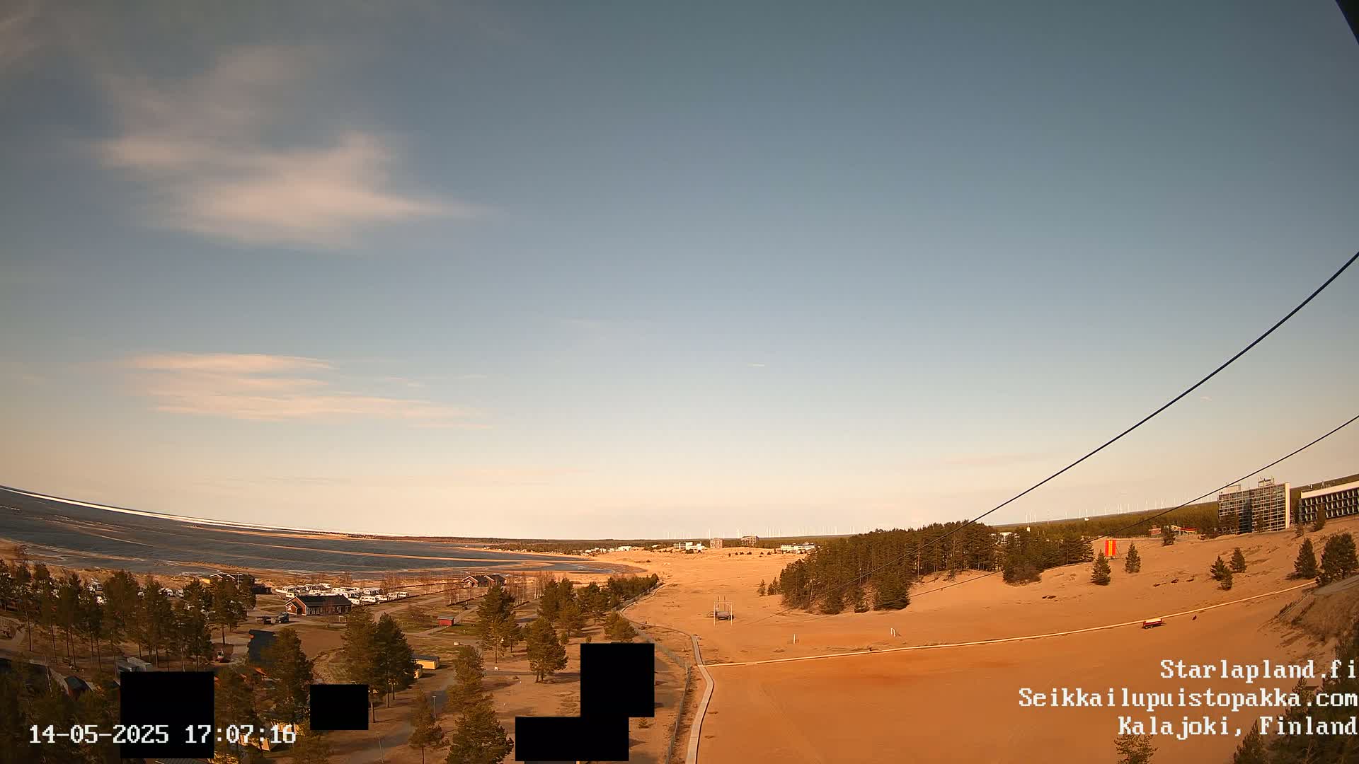 A high-angle view of a sandy landscape with sparse vegetation, a body of water in the distance, and a few buildings under a mostly clear, pale blue sky.