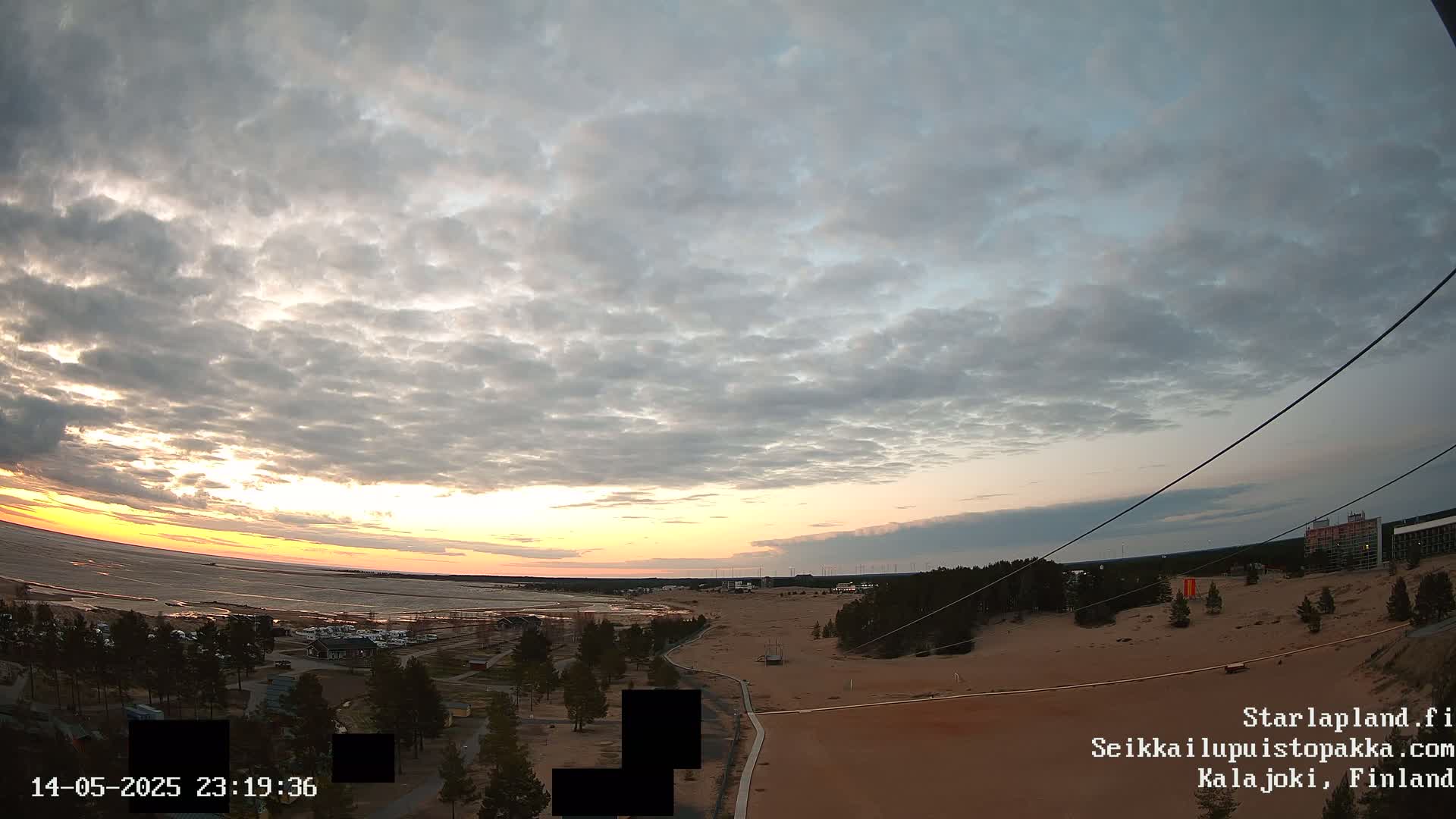 A partly cloudy sunset over a sandy landscape with a body of water in the distance, and sparse vegetation.