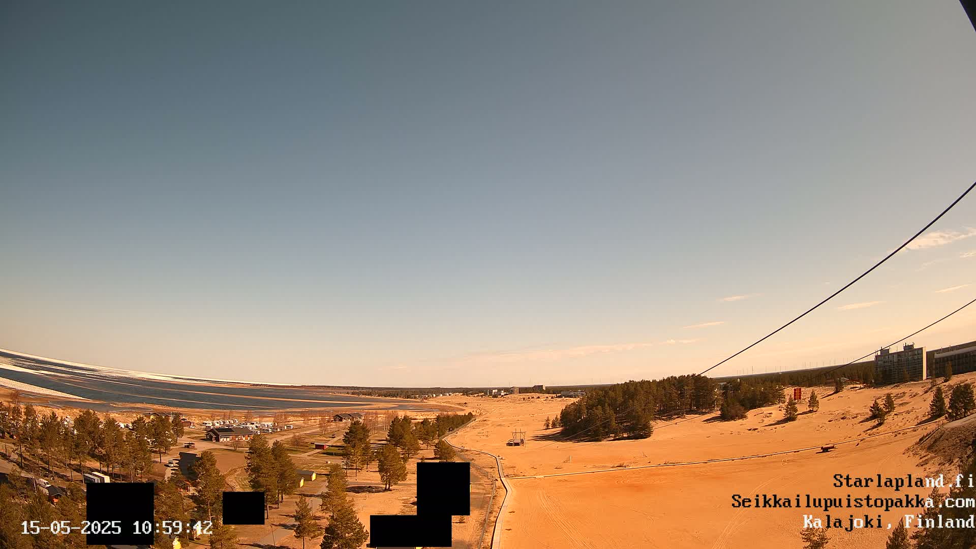 A mostly clear, sunny day reveals a sandy landscape with sparse vegetation, a body of water with some remaining ice, and a few buildings in the distance.